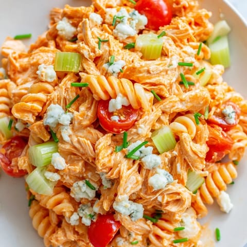 A bowl of chilled Buffalo Chicken Pasta Salad with rotini pasta, shredded buffalo chicken, diced red bell pepper, celery, cherry tomatoes, and crumbled blue cheese, drizzled with creamy ranch dressing.