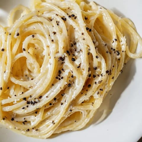 Golden strands of Spaghetti Cacio e Pepe shine in a shallow white bowl, garnished with extra Pecorino and a glass of crisp white wine nearby.